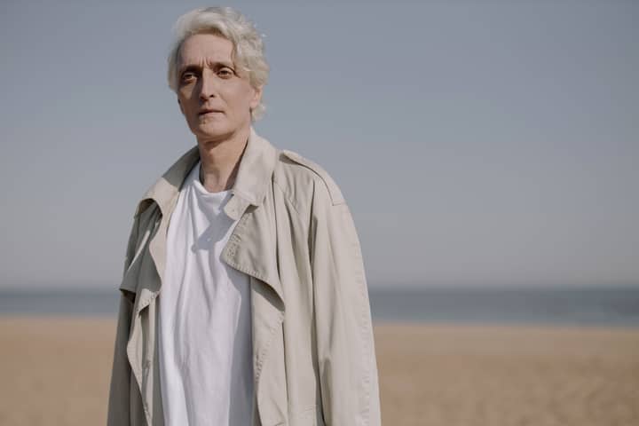 Elderly man in a coat standing on a serene beach under clear skies.