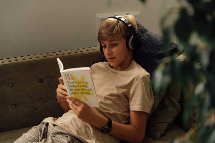 A teenager relaxes on a sofa, reading a book while wearing wireless headphones.