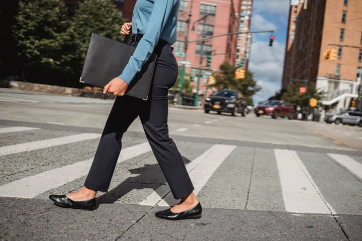 Woman in business attire crossing a street in an urban environment with buildings in the background.