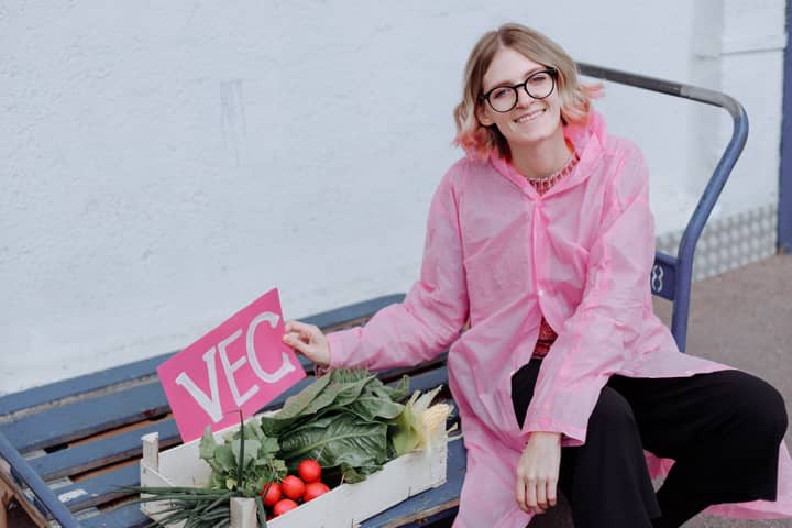A cheerful woman in a pink raincoat with fresh vegetables and a VEC sign on a wooden tray.