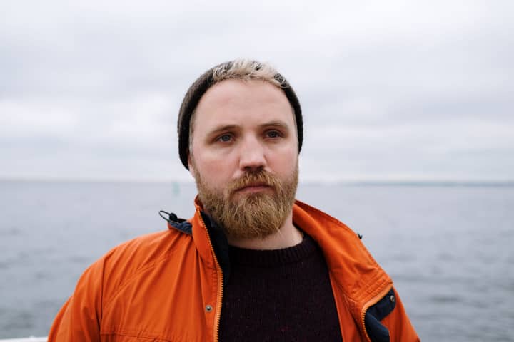 Portrait of a man wearing an orange jacket, standing by the sea with a beard and beanie.