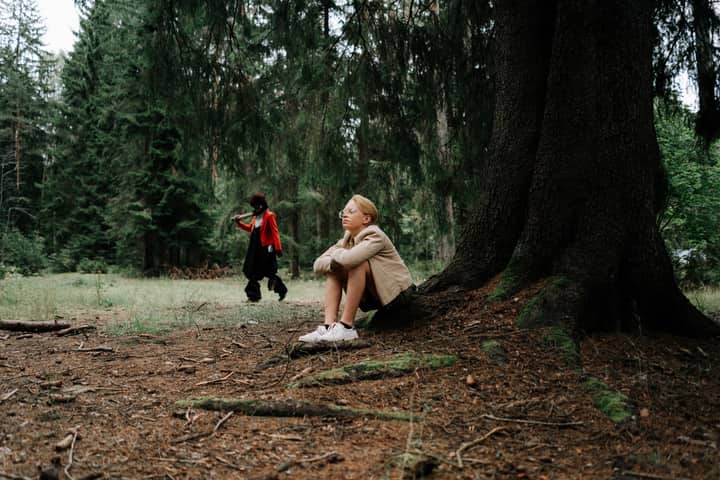 A teenager sits by a tree in a forest, unaware of a masked figure approaching.