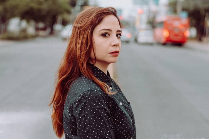 Side view of attractive young female in casual outfit with red long hair standing on road and looking at camera