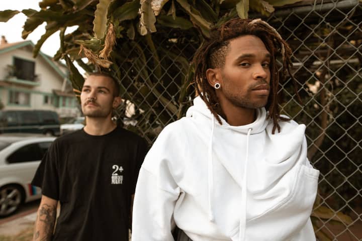 Two stylish young men posing together by a fence in an urban setting during the day.