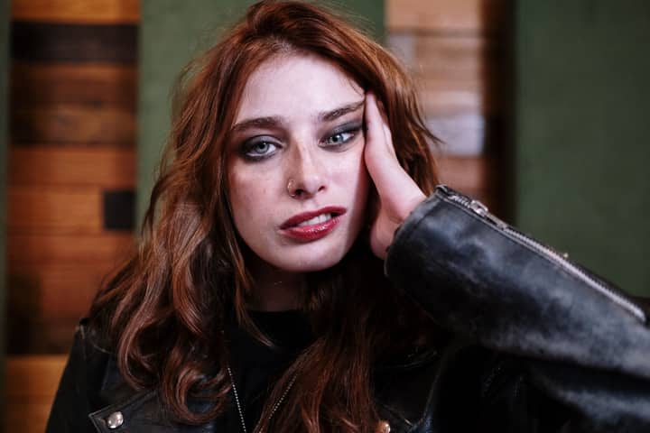 Portrait of a woman with messy hair and bold makeup, wearing a black leather jacket indoors.