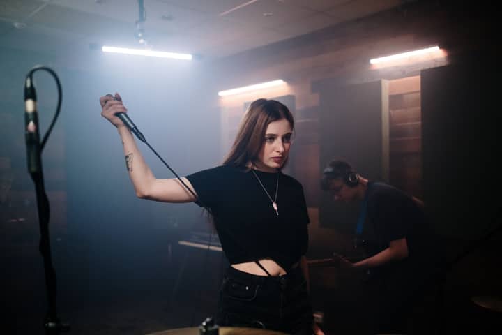 A female singer holding a microphone in a dimly lit recording studio with band.