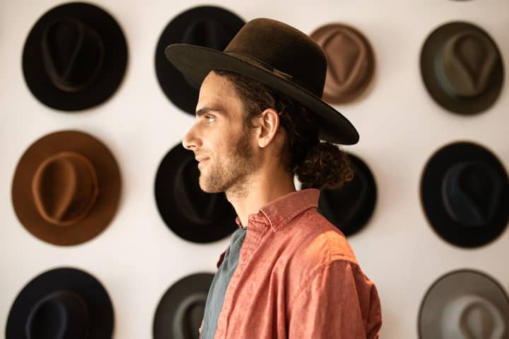 A man in a fedora stands in profile against a wall showcasing various hats.