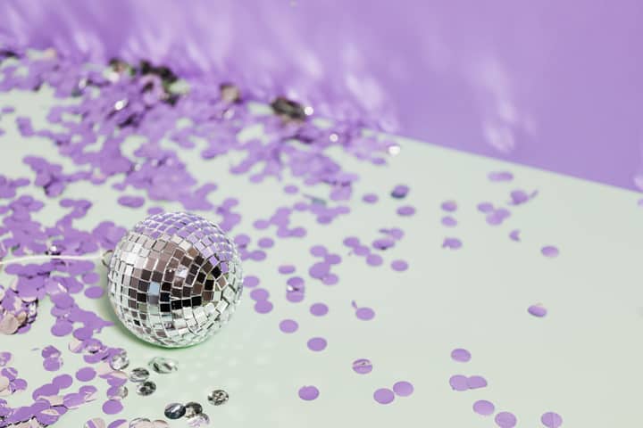 A shiny disco ball surrounded by purple confetti on a pastel background, evoking a festive mood.