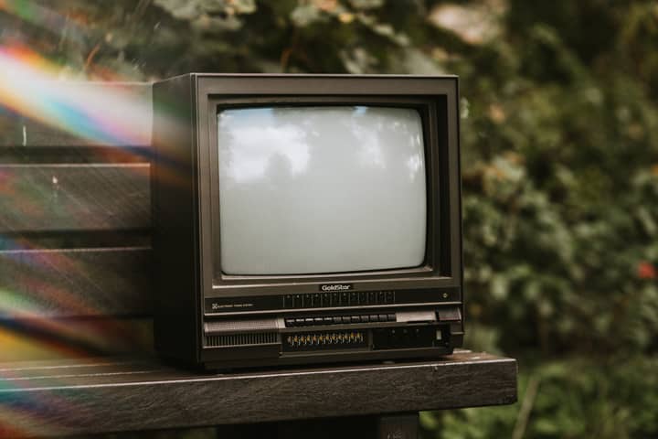 Small black old fashioned television placed on wooden bench on street with green bushes on blurred background with lens flare
