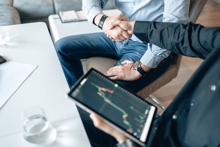 Business professionals shaking hands with a tablet displaying financial graphs.