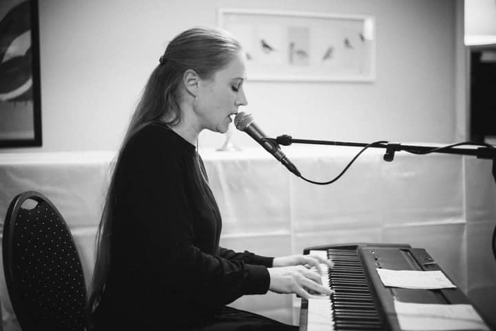 Black and white photo of a woman singing and playing the piano indoors.