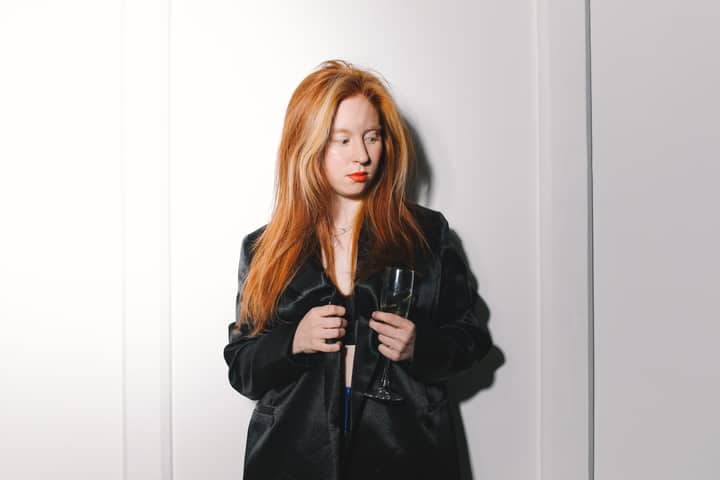 A stylish woman with red hair poses gracefully in a black jacket holding a wine glass.