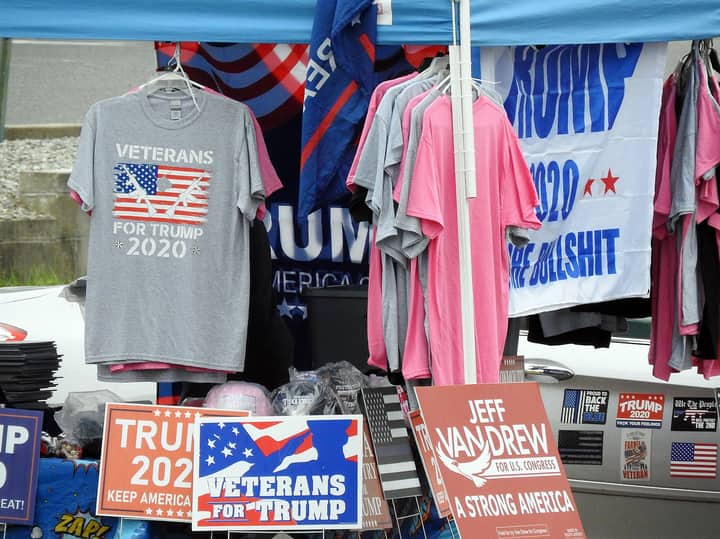 A stall displaying Trump 2020 merchandise including shirts and signs at an outdoor market.