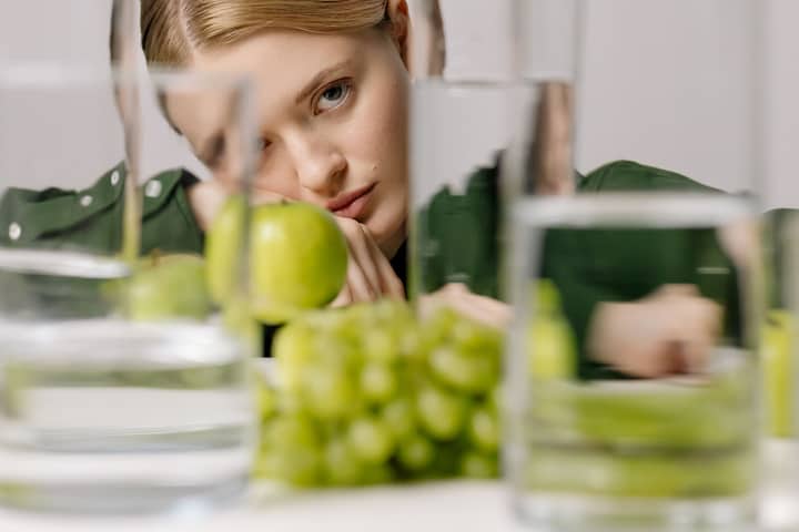 A woman in green gazing thoughtfully through glasses of water with green apples and grapes.