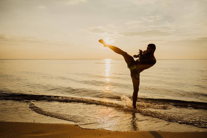 Martial artist performing a high kick on a beach during sunset, creating a dynamic scene.