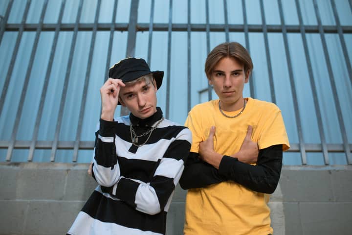 Two trendy teenagers posing in casual outfits against a metal fence background.