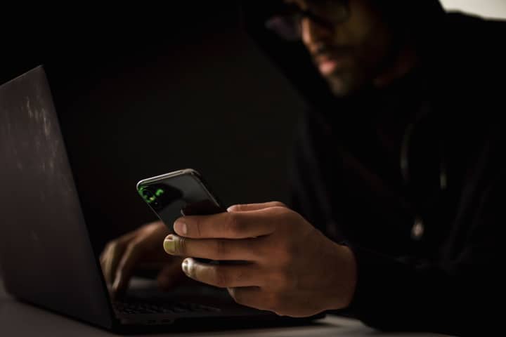 Crop anonymous ethnic male cyber spy with cellphone and netbook hacking system in evening