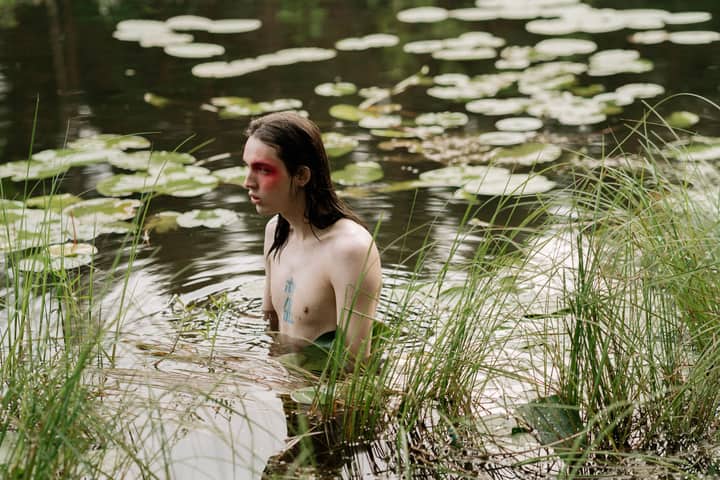 A shirtless man with painted face standing in a serene lake surrounded by nature.