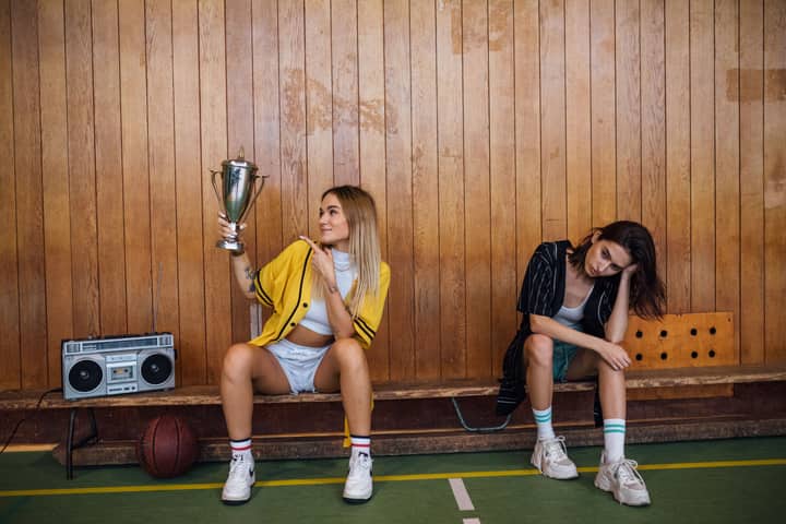 Two young women on a bench, one celebrating with a trophy, depicting competition.