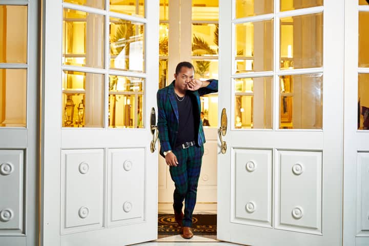 A stylish man in a plaid suit stands confidently at a luxurious entrance with open doors.