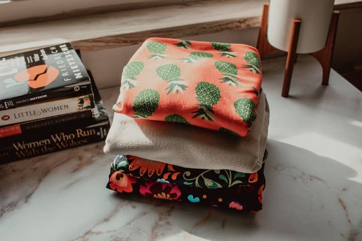 Stacked colorful patterned fabrics beside popular books on a sunlit desk.
