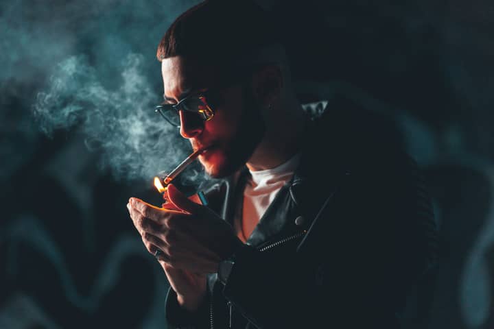 Close-up of a man in sunglasses lighting a cigarette in a dark setting, exuding a moody vibe.
