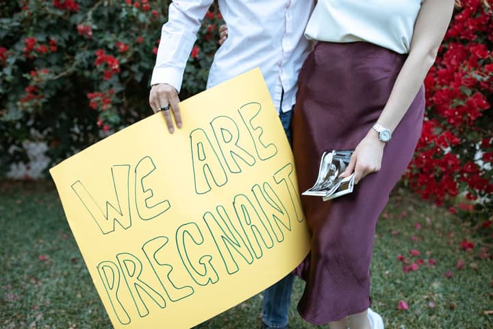 A couple holds a yellow sign saying 'We Are Pregnant' outdoors with red flowers in the background.