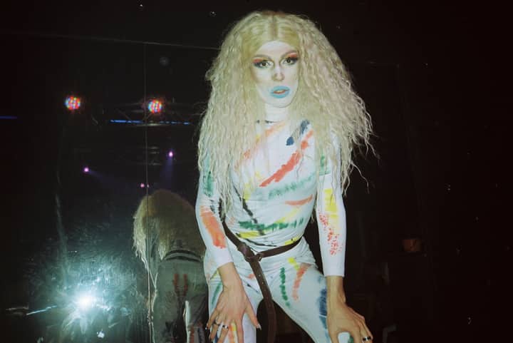 A drag queen in colorful costume posing with reflection on stage.