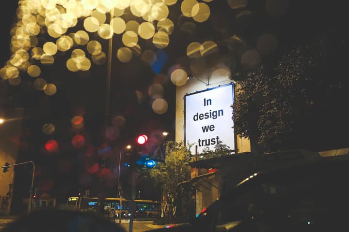 A city street at night featuring an illuminated billboard with the phrase 'In design we trust.'