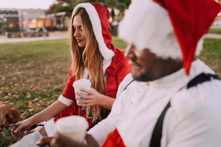 Couple in Santa hats enjoying coffee outdoors, celebrating holiday season in a park.