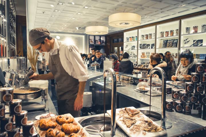 Chef prepares baked goods in a bustling bakery shop with customers.