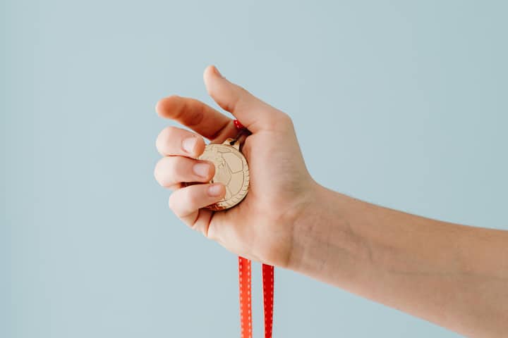 A close-up image of a hand holding a gold medal with a red ribbon against a blue background.