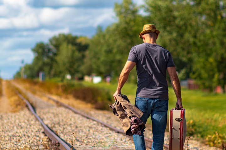 Man walking down railroad tracks holding a suitcase and coat on a sunny day.