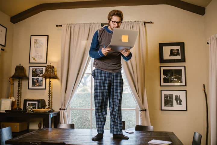 Man in pajamas standing on table with a laptop indoors, focused on remote work or study.
