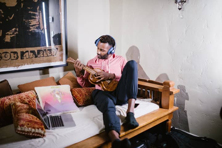 Man in pink hoodie playing ukulele on a couch, wearing headphones, with a laptop nearby.