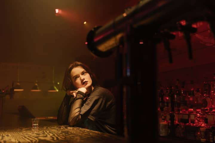A woman in a dimly lit bar, seated at the counter with a contemplative look.