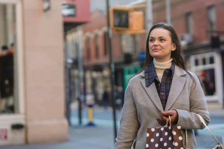 Dreamy young woman in warm coat and gloves with gift packages and purse strolling in street near buildings and road while looking away in town in daylight