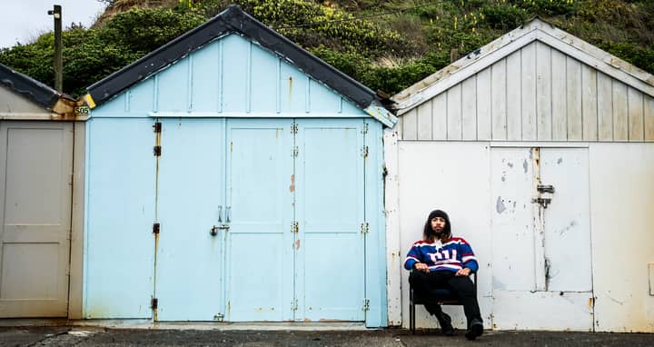 Man sitting on chair beside colorful beach huts in Porirua, New Zealand.