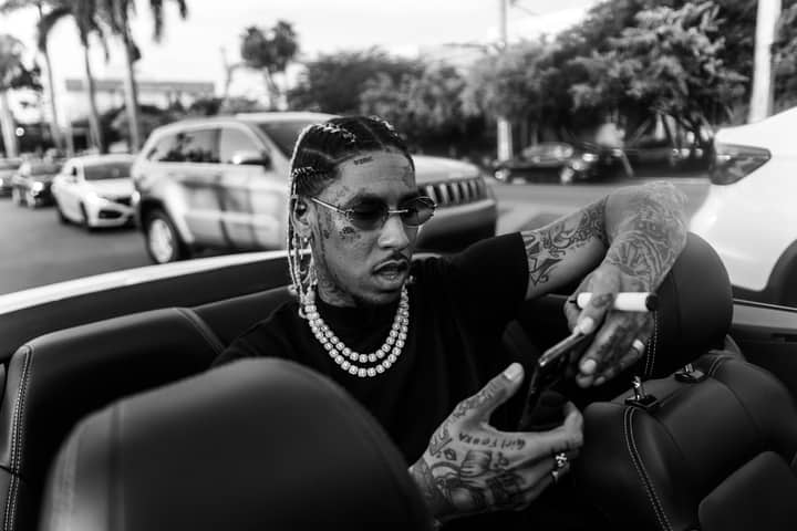 Black and white of African American tattooed male looking at mobile phone in sunglasses with dreadlocks and beads on neck