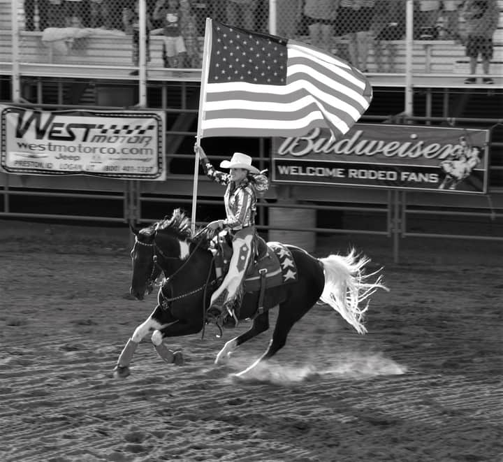 A cowgirl rides a horse with the American flag at a rodeo event. Black and white action shot.