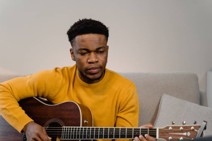 A man in a yellow sweater plays an acoustic guitar while sitting in a cozy indoor setting.