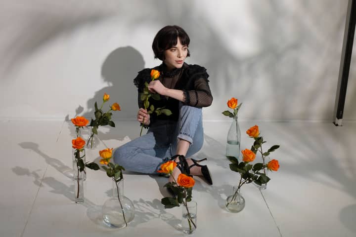 Full length of young dreamy female in stylish outfit sitting on tile floor with glass vases with orange roses in water near white wall with shades while looking away