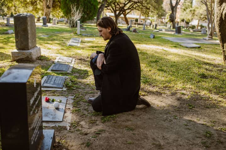 A man kneeling at a gravestone in a cemetery, holding a red rose during an outdoor visit.