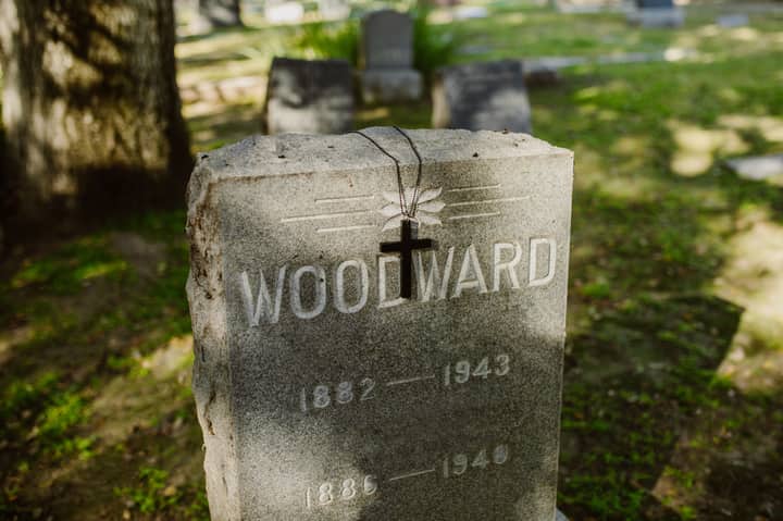 A peaceful cemetery scene with a gravestone marked 'Woodward' and a cross necklace hanging in sunlight.