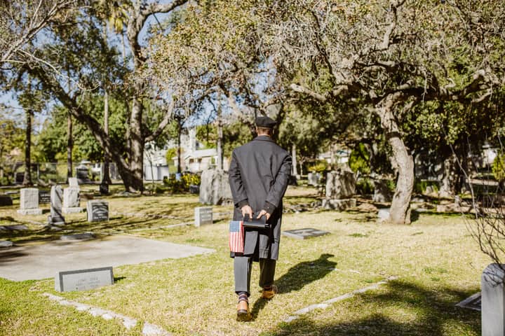 A man in uniform holds a flag, walking through a sunlit cemetery, evoking remembrance.
