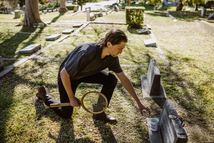 A man kneels with a tennis racket by a tombstone, symbolizing grief and remembrance.