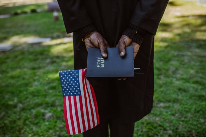Close-up of hands holding a Holy Bible and American flag in a green outdoor setting.