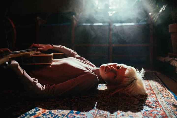 A woman with blonde hair lying on a patterned rug, holding a guitar in an artistic setting.
