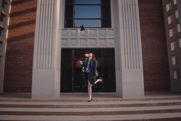 Young woman celebrating her graduation by tossing cap outside the university building.