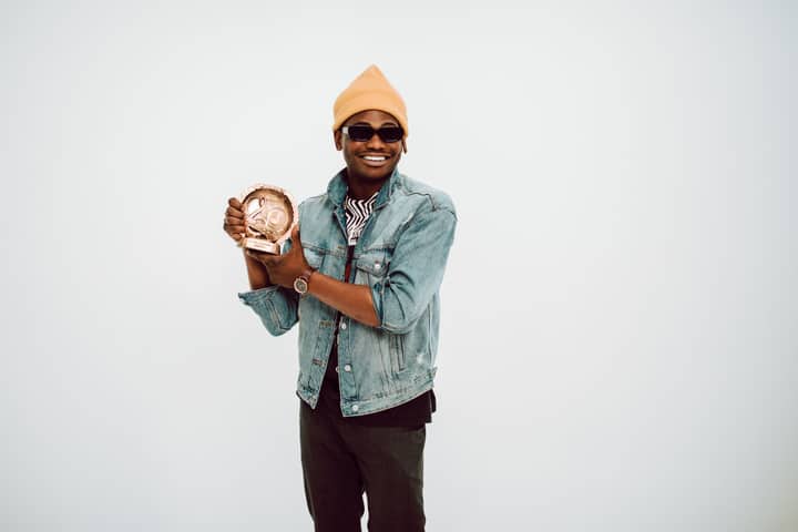 Smiling man in casual attire with sunglasses holding a trophy in a studio environment.
